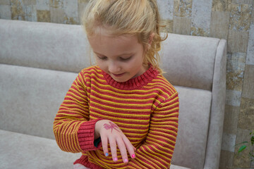 Fototapeta premium Cute little blonde girl looking at her glitter tattoo on hand ,a blonde girl in a striped sweater showing a colorful manicure and a temporary tattoo in the shape of a butterfly,
