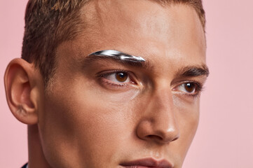 Obraz premium Chrome liquid metallic pigment coats the right eyebrow in a thick, reflective sculptural stroke against pale skin and a matte dusty pink backdrop. Sharp specular highlights dance across the silver