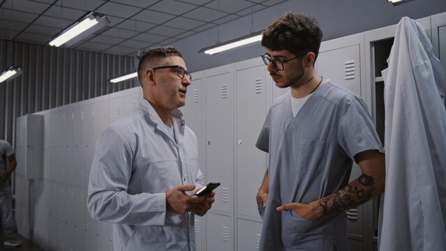 After Busy Workday, Two Male Doctors Relax by Grey Metal Lockers, Viewing Sport News on Smartphone and Chatting About Personal Interests in Locker Room. Concept of Shift End and Employee Wellness.