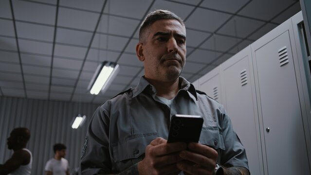 Serious Middle Aged Male Security Officer With Prominent Arm Tattoos Stands by Grey Metal Locker, Concentratedly Typing Message or Email Response on Smartphone After Finishing Airport Security Shift.