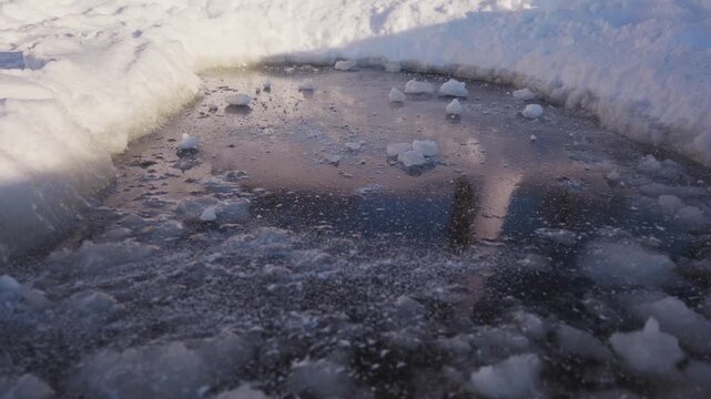 Broken chunks of hard snow are scattered across a dark frozen ice patch between white snowbanks, showing rough winter texture, icy ground, and cold outdoor surface details. High quality 4k footage