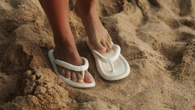 Sand covered sandals beside bare sunlit feet. Closeup of white flipflops on warm golden sand as a beachgoer tests footwear, shifting toes, leaving soft footprints intimate, relaxed summer atmosphere