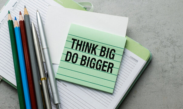 Green sticky note reads Think Big Do Bigger. Pens and pencils rest beside open notebook. Focused, motivational message for ambitious goals.