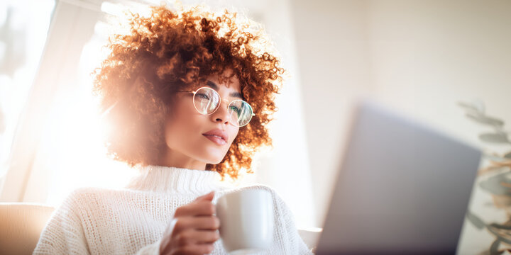 Pensive woman with curly red hair and glasses holding coffee by window