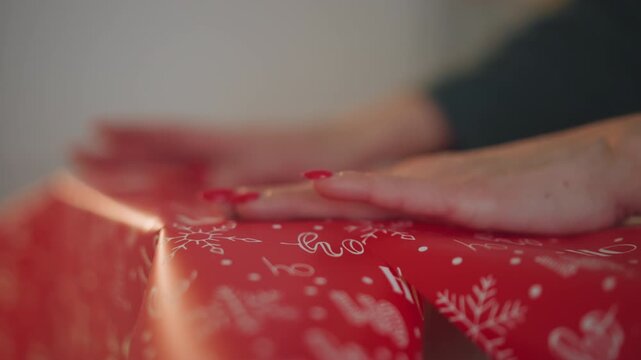 Closeup hands wrapping christmas present in red patterned paper, careful creasing and smoothing, manicured nails, soft warm light, cozy living room atmosphere, intimate holiday preparation, subtle