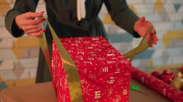 Hands wrapping red christmas present on table with green ribbon and gold tape, holiday ornaments and scissors nearby, DIY crafter ties neat bow in warm cozy indoor mood with patterned wall backdrop.