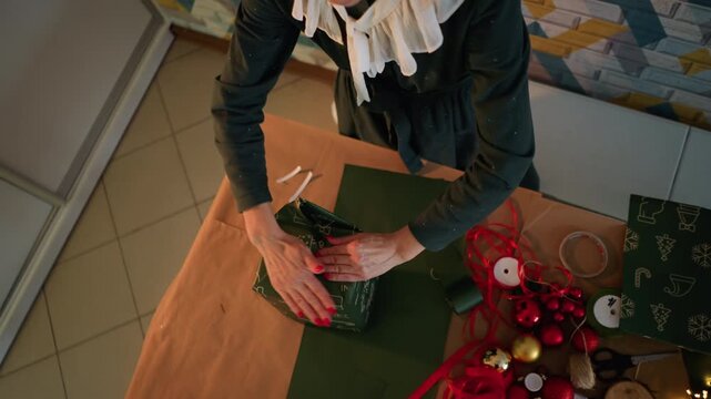Blonde white woman wrapping christmas present on wooden table, arranging ribbon and ornaments, tying neat bow and smoothing green patterned paper, warm ambient light and festive red baubles, tiled