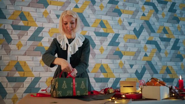 Woman wrapping christmas present on table, cozy home setting with patterned brick wall, red ribbon, scissors, lit candles, string lights, gift boxes, oranges, warm lighting, joyful holiday mood,