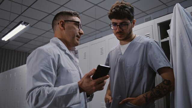 Senior Surgeon Mentors Younger Intern With Tattoos, Using Smartphone to Review Interns Scientific Research Paper and Findings in Staff Changing Area. Medical Education and Mentorship Concept.
