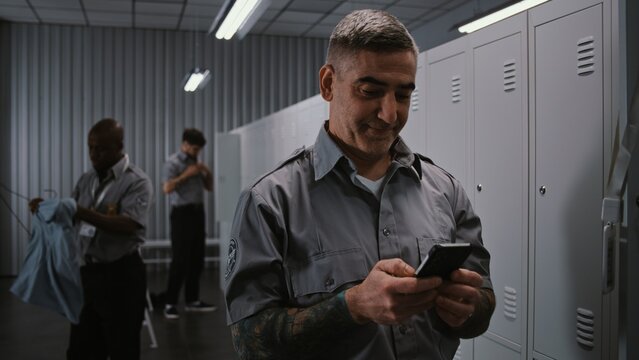 Muscular Male Security Officer With Tattoos at Locker Hangs Up ID, and Uses Smartphone to View Notifications and Social Media After Long Airport Security Shift. Workplace Communication Concept.