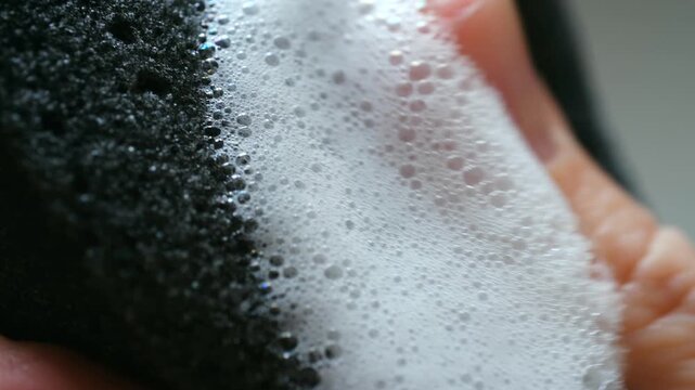 Detailed macro shot of hand spreading white soapy foam onto black porous sponge, with the suds and bubbles slowly absorbing into the surface, illustrating cleaning and hygiene concepts