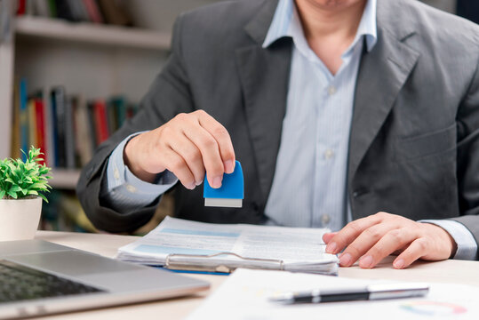 Businessman hand using blue rubber stamp to approve contract document on desk. Corporate executive stamping official paper for legal certification. Notary public authority concept.