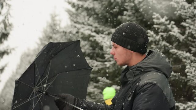 Caucasian man black umbrella in snow, standing on pinelined road with protective posture, hooded coat and gloved hand, subtle forward gaze, cinematic muted tones, passerby with neon backpack