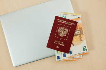 Fototapeta premium Laptop with Russian passport and 200 and 50 euro banknotes on wooden desk for immigration and travel planning concept. Online booking, relocation budget and digital vacation preparation.