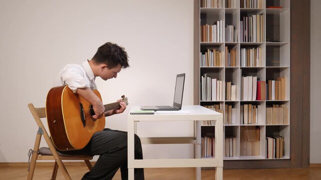 Man playing acoustic guitar while participating in an online lesson, looking at a laptop computer on a table in his home office with a bookshelf background