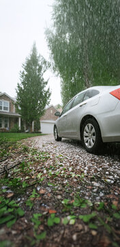 Hail damaged silver vehicle in a driveway, suburban home parked in front of house. A severe storm, low angle, hail stones, tree debris  on the ground, hail-damaged roof, car weather, vertical. mobile
