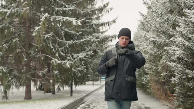 Man adjusting backpack on snowy path, tightening gloves and pulling beanie while walking past frosted pine trees, breath visible in cold air, wearing black parka and jeans, urban park walkway