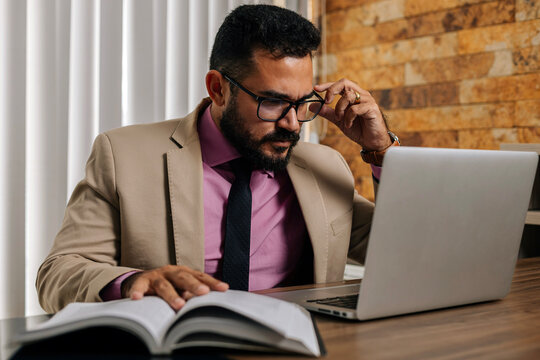 Businessman reading a book while working on laptop at desk, studying and analyzing information in a professional office setting.