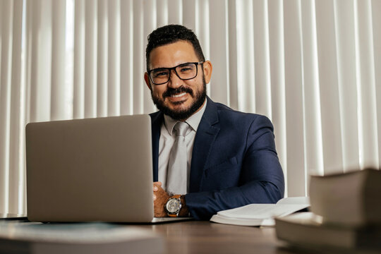 Smiling businessman using laptop at desk, working online and managing tasks in a modern and comfortable office environment.