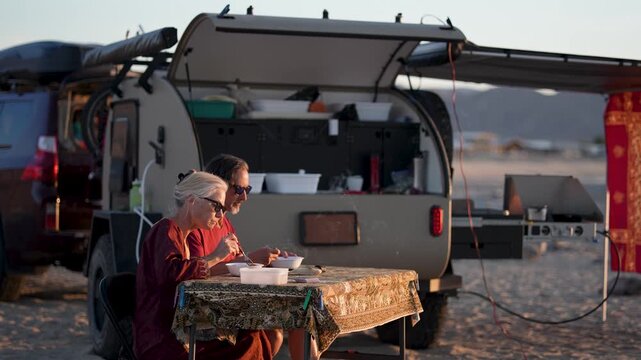 Mature man and woman enjoy dinner while camping at the beach with their teardrop trailer as the sun sets in Baja California Mexico.