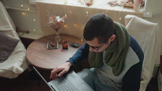 Caucasian man working on laptop at round table wearing sunglasses and green scarf, adjusting screen and typing, focused remote work vibe, fairy lights and dried flowers in background, home office