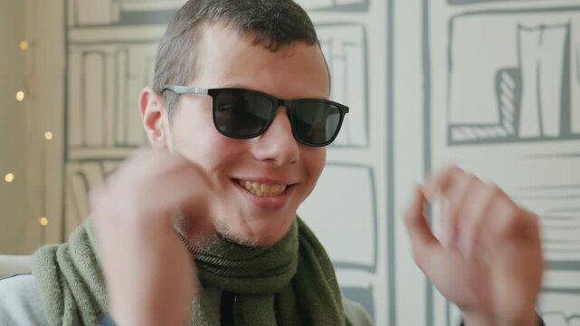 Young man dons sunglasses against illustrated library backdrop. Studiostyle portrait with sketchbook wall and string lights, scarf accessory and confident attitude shifting to a relaxed laugh,