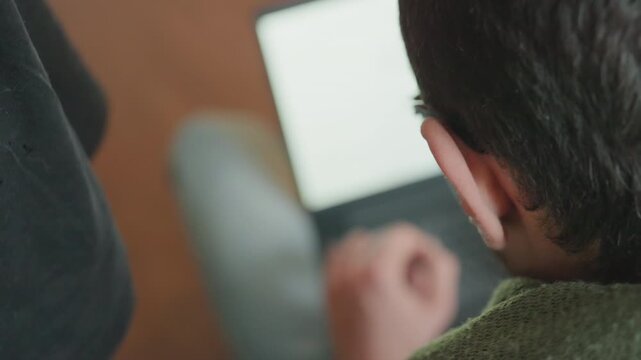 Overshoulder caucasian student typing on laptop, closeup of hands and shoulder with soft screen glow, blurred background bokeh, focused posture and intent expression, university assignment research