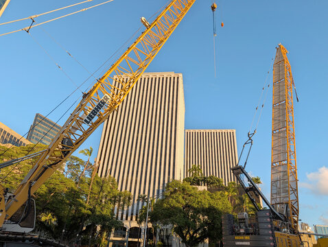 Construction work occurs in Oahu near tall buildings as cranes lift materials during the day