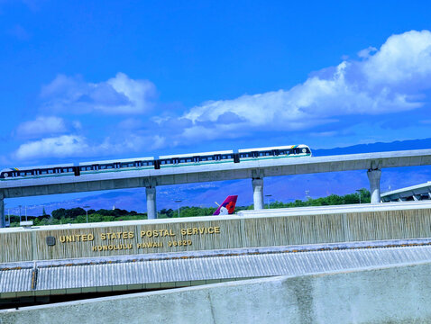 A Skyline train travels above the United States Postal Service building at the airport in Honolulu, Hawaii. The sky is clear with some clouds.