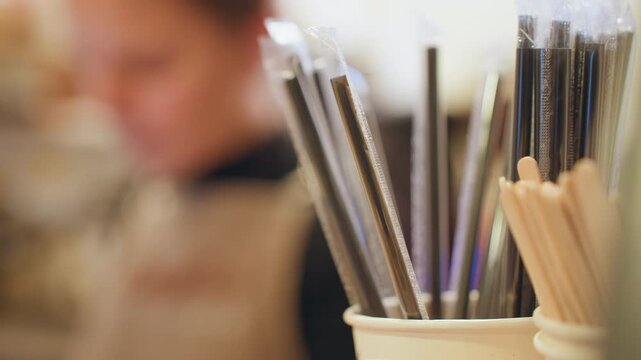 Blurred barista behind stirrers and straws in cup, busy cafe backdrop, paper cup holder, motion suggestion, shallow focus on utensils, warm ambient lighting, candid counter service scene.