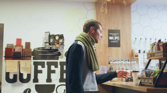 Caucasian man at coffee counter smiling green scarf paying at register barista preparing espresso jars and syrups on shelves warm bistro lighting casual winter layers takeaway cup in hand bright