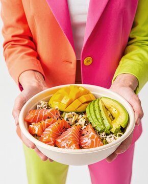 Person in colorful outfit holding a refreshing salmon, avocado, and mango poke bowl. Perfect for health-conscious blogs or magazine features.