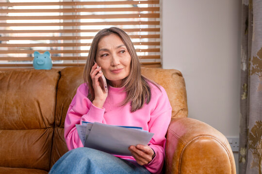 Woman sitting on a couch talks on phone while holding papers in her hand during morning hours at home 
