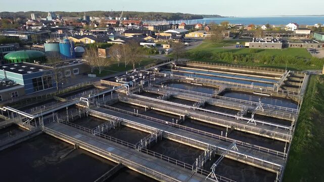 Wastewater treatment plant with multiple filtration basins, processing tanks and interconnected purification systems. The facility is located near a coastal town, highlighting essential water treatmen