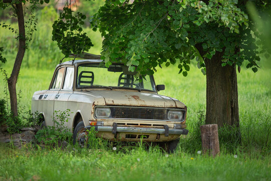 Abandoned Rusty Vintage Car Under Tree In Overgrown Green Field Evoking Rural Decay