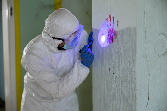Forensic specialist in a hazmat suit searching for biological evidence at a crime scene using an ultraviolet flashlight