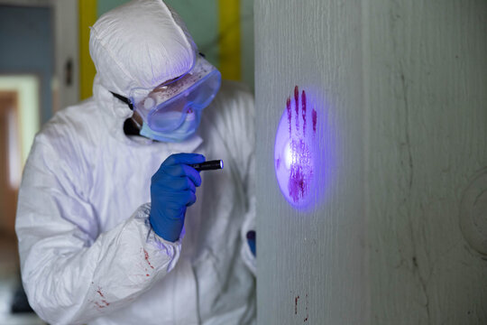Forensics officer in hazmat suit illuminating a gory handprint on a wall with ultraviolet light, collecting critical evidence