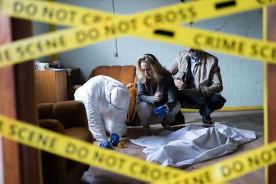 Forensic team examining evidence and a body covered with a sheet, behind do not cross police tape at a crime scene