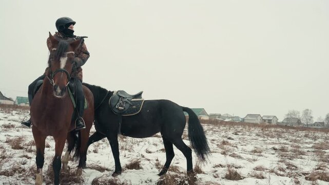 Caucasian rider on brown horse snowy field tethered black mare nearby heavy coat and helmet overcast sky rural village background patient equine companion grazing frost covered grass leather saddle