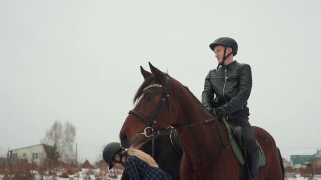 Rider scans village. Caucasian rider in helmet watches serene winter village scene attentively. Man dressed in helmet atop chestnut horse surveys calm village environment amidst winter surroundings