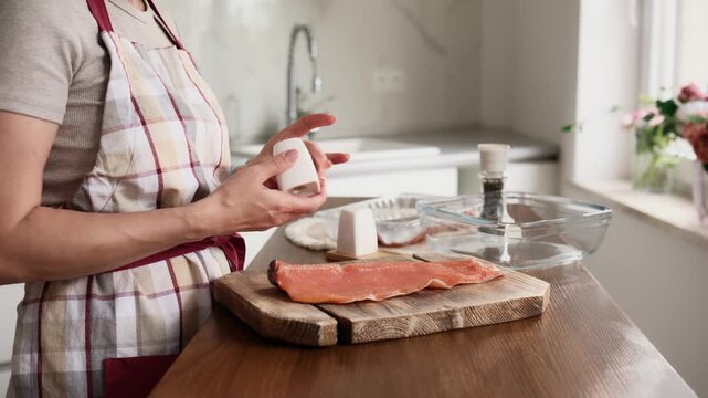 Woman prepares salmon on cutting board. She sprinkles seasoning on the raw fish in a bright kitchen.