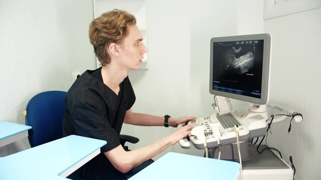 Young male caucasian sonographer operating ultrasound console, analyzing abdominal scan on grayscale monitor in bright clinic room. Focused posture at workstation, medical equipment, sensor and cables