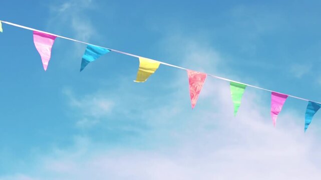 Colorful triangular flags in vibrant shades of pink, blue, yellow, and green sway gently against a bright blue sky with soft clouds, creating a festive atmosphere
