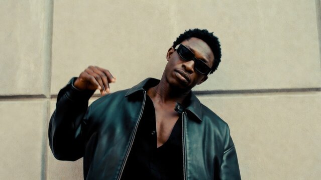 Man wearing leather jacket posing with sunglasses by wall
