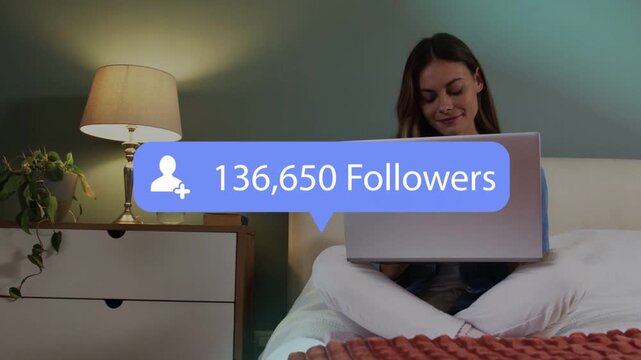 Woman sitting crosslegged on bed using laptop while social follower overlay updating causing smile