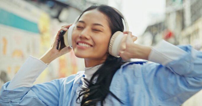 Happy young Asian woman with closed eyes smiles and sways gently to her favorite music. She wears white wireless headphones and holds a smartphone on a city street.