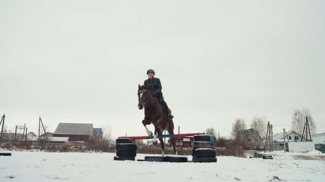 Student on horse at jump training, instructor observing from distance near tire and pole obstacle, measured posture and steady reins, snowy open arena and farmhouse backdrop, concentrated education