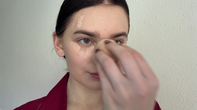 Young woman applying creamy foundation, blending liquid concealer with makeup sponge, preparing smooth skin for daily glamour routine indoors, demonstrating cosmetic beauty application process.