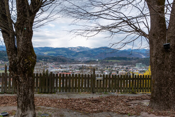 Obraz premium Baernbach, Austria – March 28, 2026: A panoramic view over a small town with houses, industrial buildings and distant mountains framed by trees and a wooden fence in the foreground.