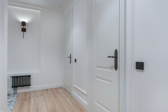 A person walks through a modern hallway with white walls and wooden flooring. There are several doors and a wall light fixture offering illumination